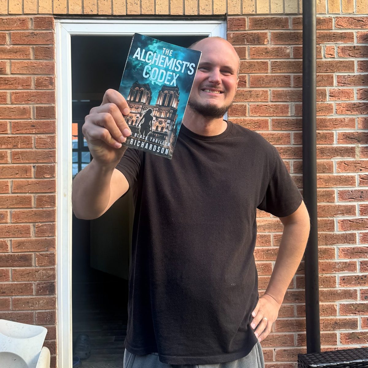 Author Luke Richardson smiling and holding up The Alchemist’s Codex paperback